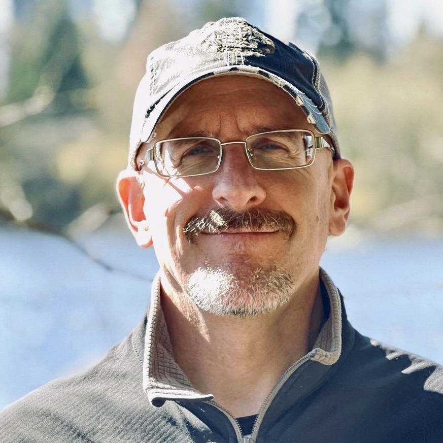 Man with moustache, glasses, and a ball cap, smiling at camera, standing outside by a lake.